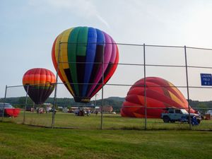 Three colorful hot air balloons at a festival, one rainbow-striped inflated and two partially inflated on green grass.