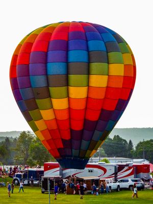 Colorful rainbow hot air balloon launching at an outdoor festival with crowds and RVs on a green field.