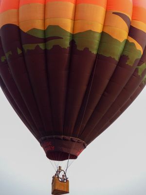 Colorful hot air balloon with mountain landscape design soaring in sky with passengers visible in wicker basket below.