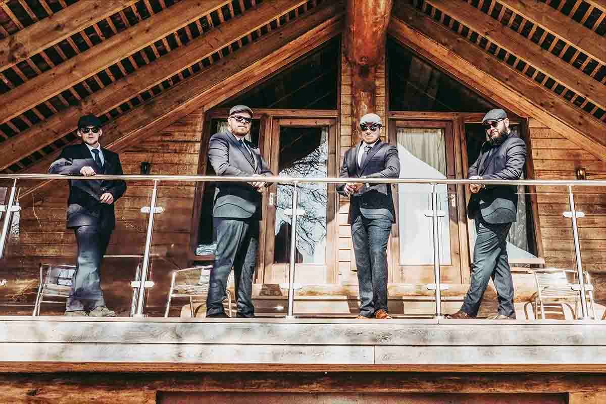 Four men in suits and sunglasses stand on a wooden chalet balcony, posing confidently in a stylish group photo.