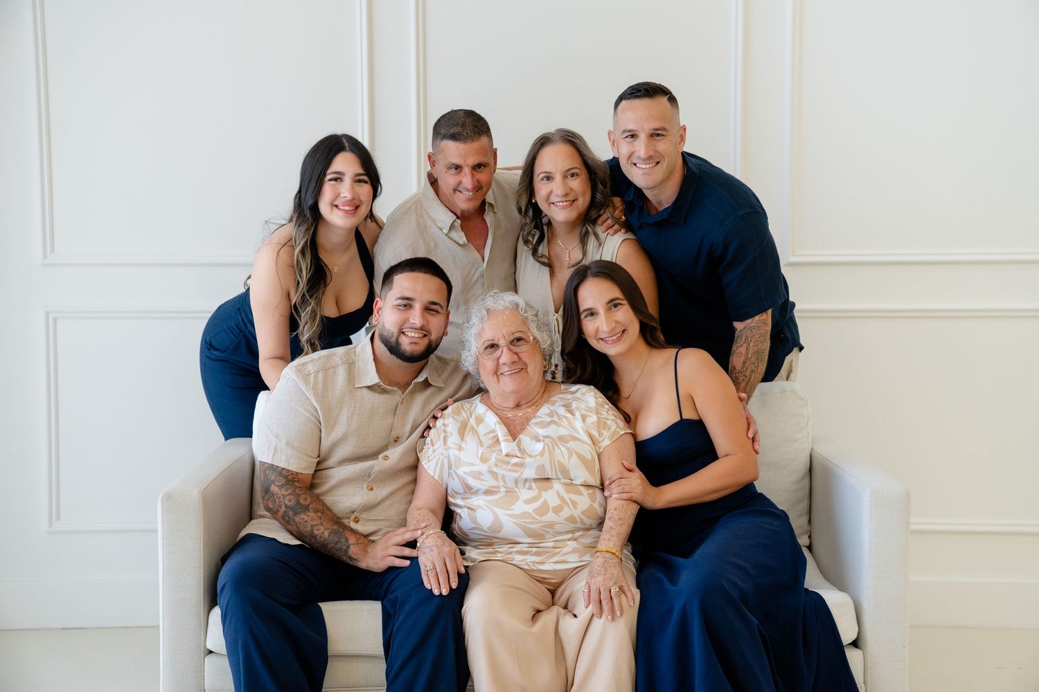 Family of seven posing together in a bright white studio, with elderly woman seated centrally surrounded by smiling loved ones.