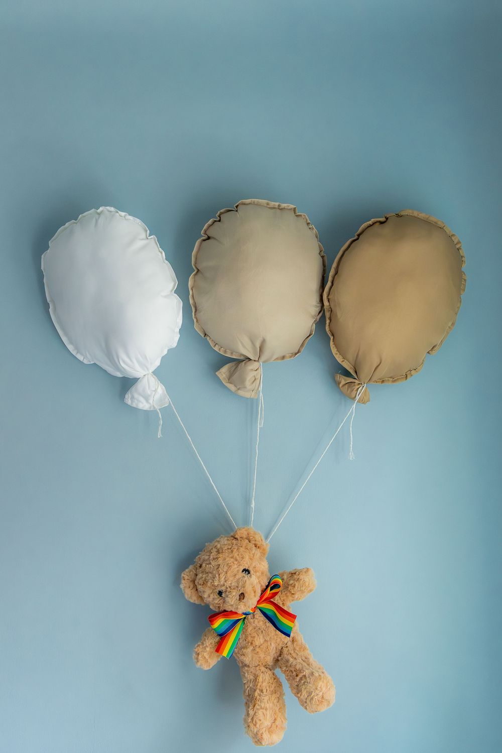 Teddy bear with rainbow bow tie floating with three fabric balloon pillows on blue background.