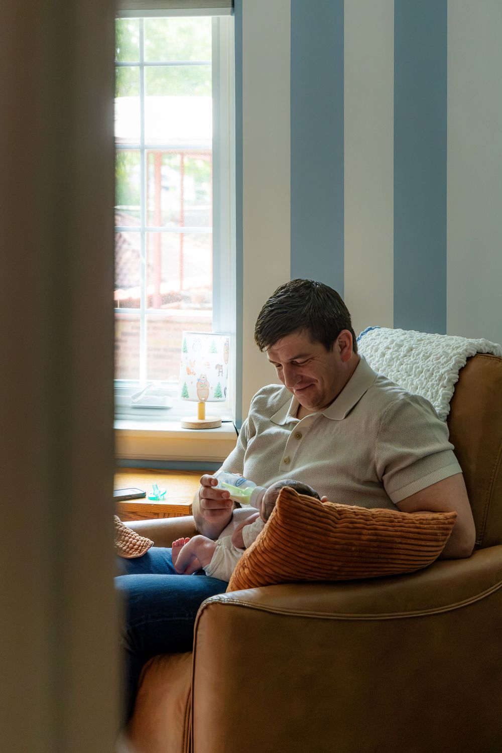 Father sitting in armchair by window holding newborn baby feet in cozy striped nursery room.