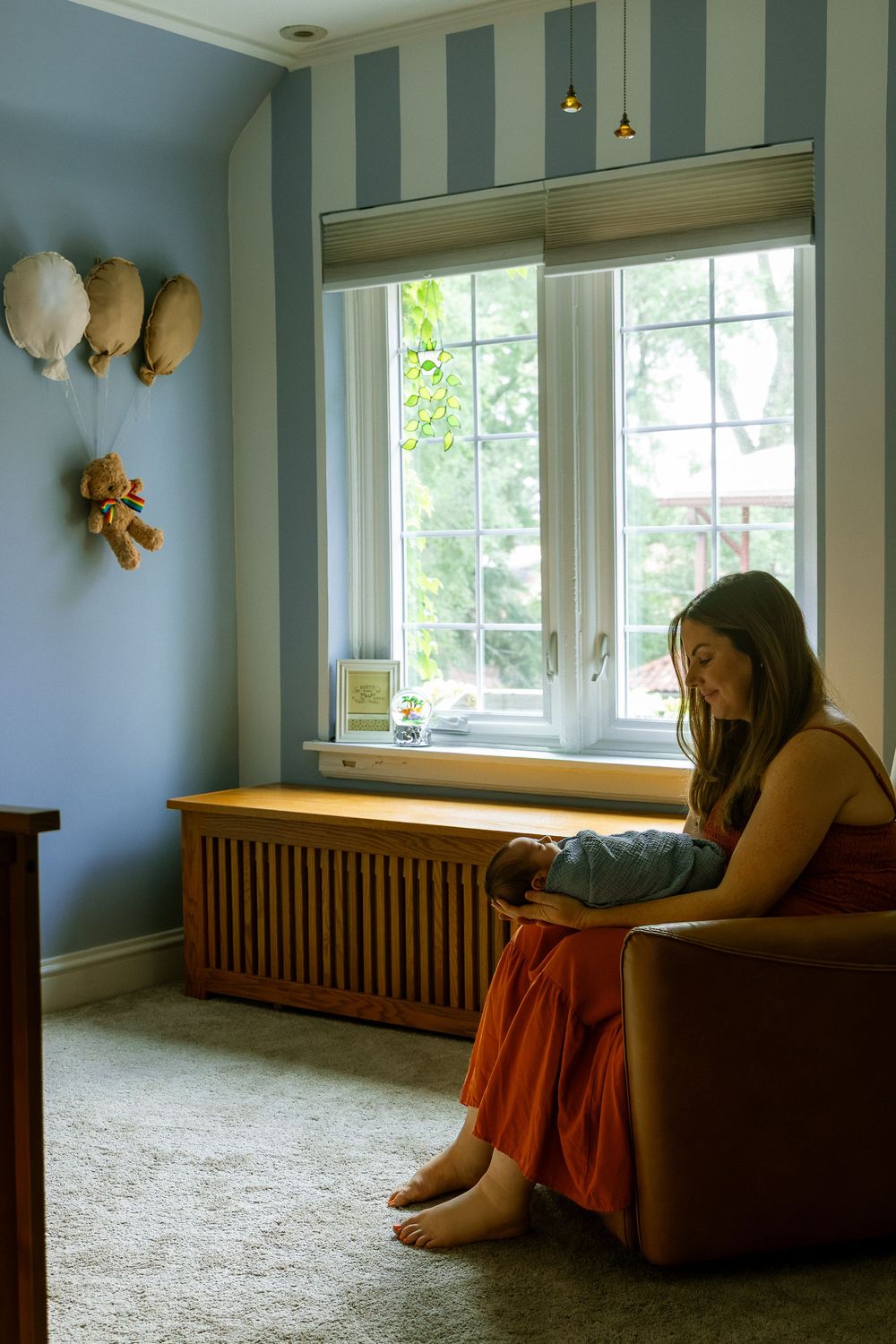 Young woman sitting barefoot in armchair holding newborn baby in a softly lit nursery with blue striped walls.