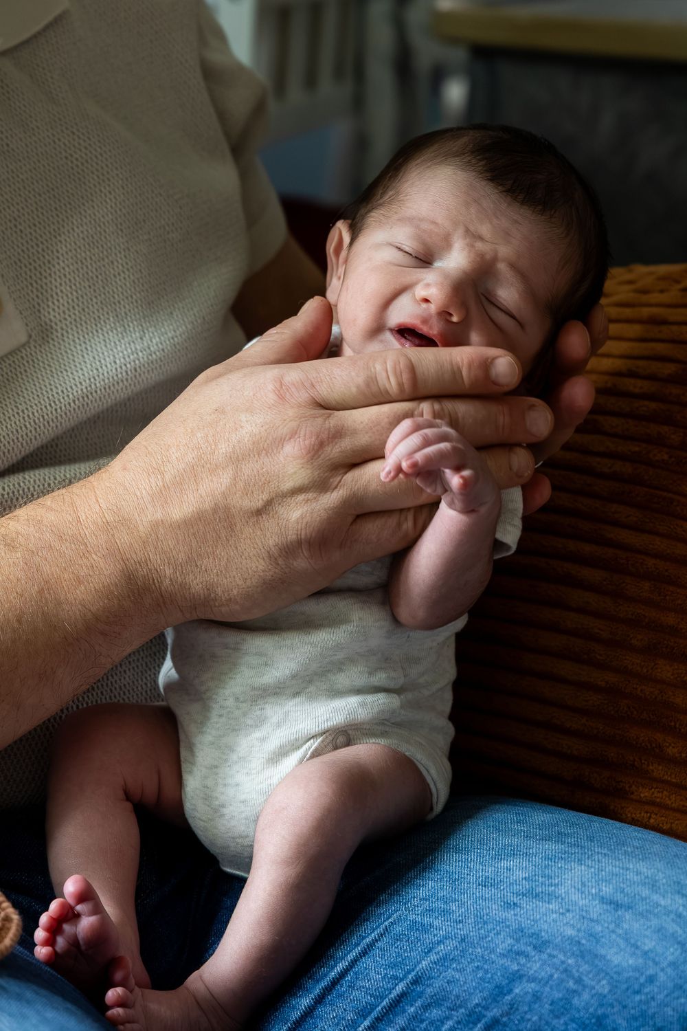 Adult hands gently cradling a crying newborn baby dressed in white, sitting on a lap in soft indoor lighting.
