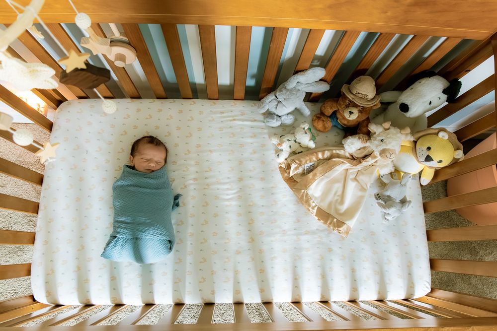Newborn baby swaddled in blue sleeping in a wooden crib beside stuffed animals on a white mattress.