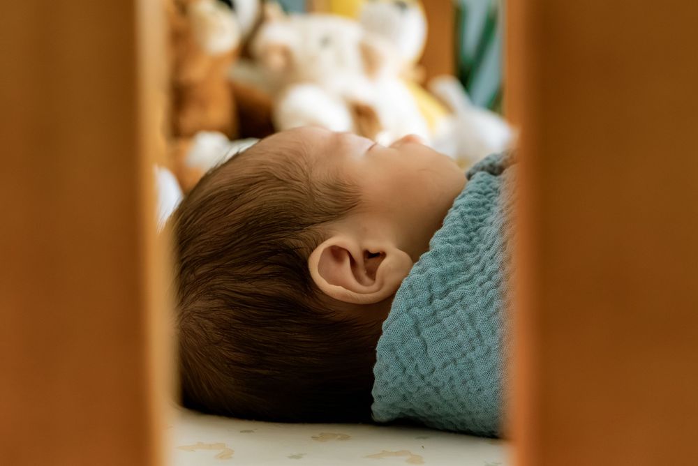 Peaceful newborn baby sleeping in a wooden crib wrapped in a soft blue blanket with stuffed animals nearby.