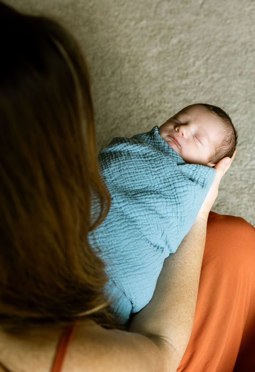 A mother in an orange dress cradles her sleeping newborn baby swaddled in a soft blue muslin wrap.