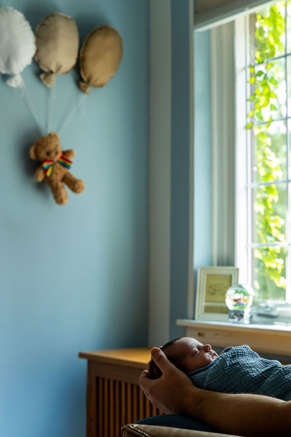 Newborn baby swaddled in blue, lying near a sunlit window in a nursery with a teddy bear balloon wall decor.