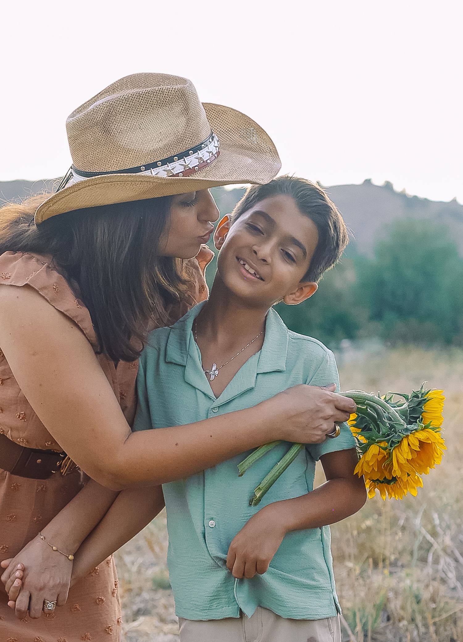 A woman in a straw hat kisses a smiling boy holding bright sunflowers in an outdoor field setting.