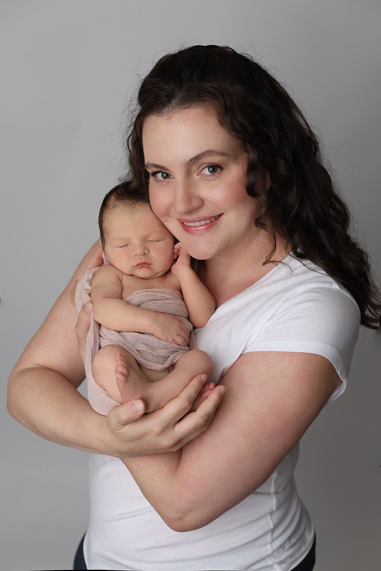 A smiling young woman in a white t-shirt tenderly holds a sleeping newborn baby wrapped in a pink swaddle.