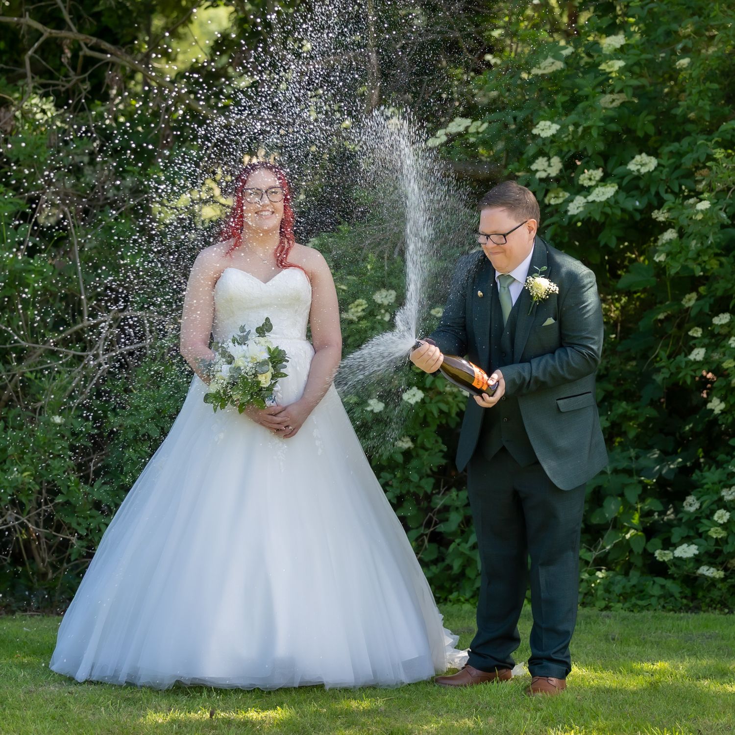 Bride in white ballgown and groom in gray suit popping champagne bottle outdoors in garden setting. Wedding, Casual Captures