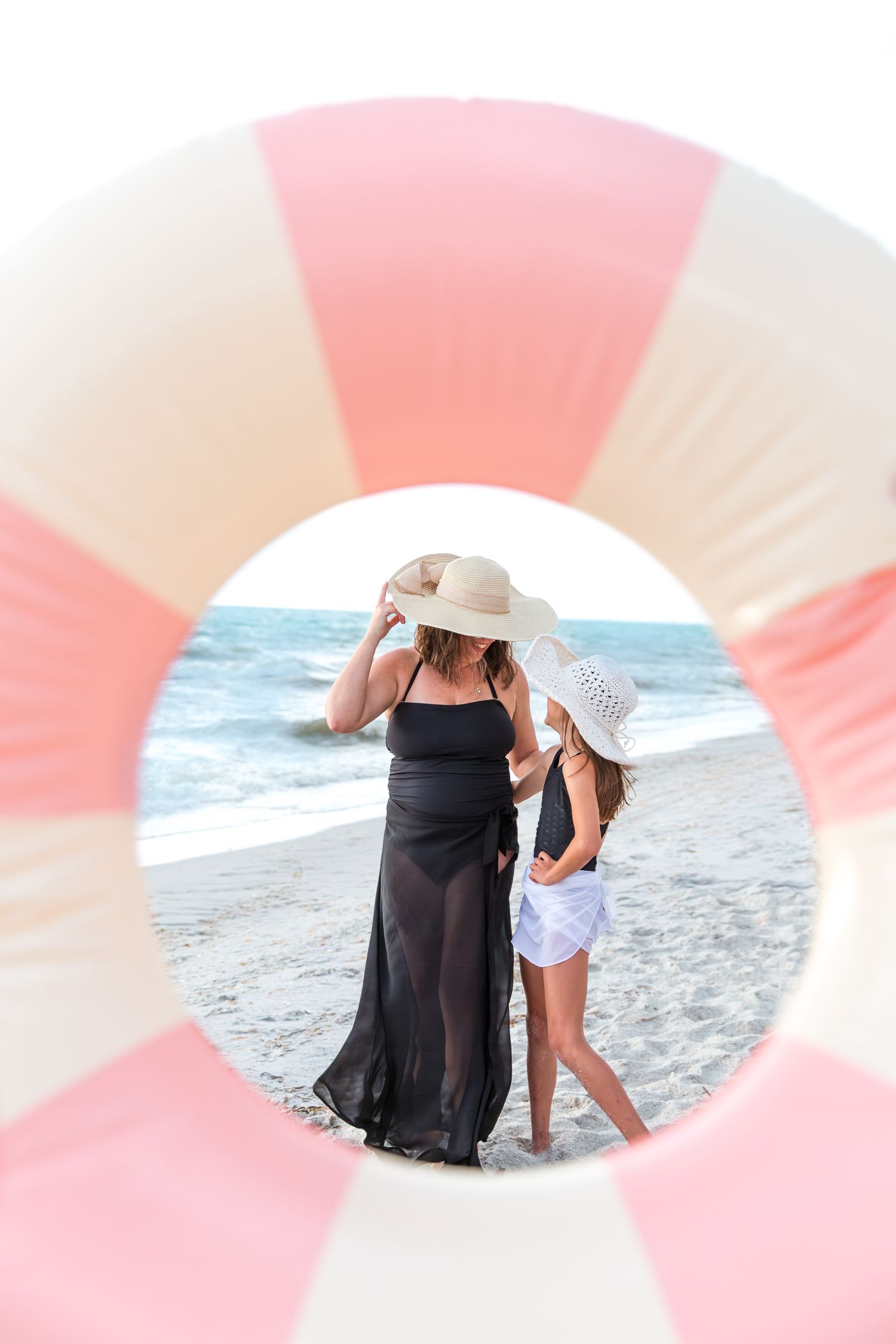 Woman and girl in sun hats viewed through pink striped pool float at beach.