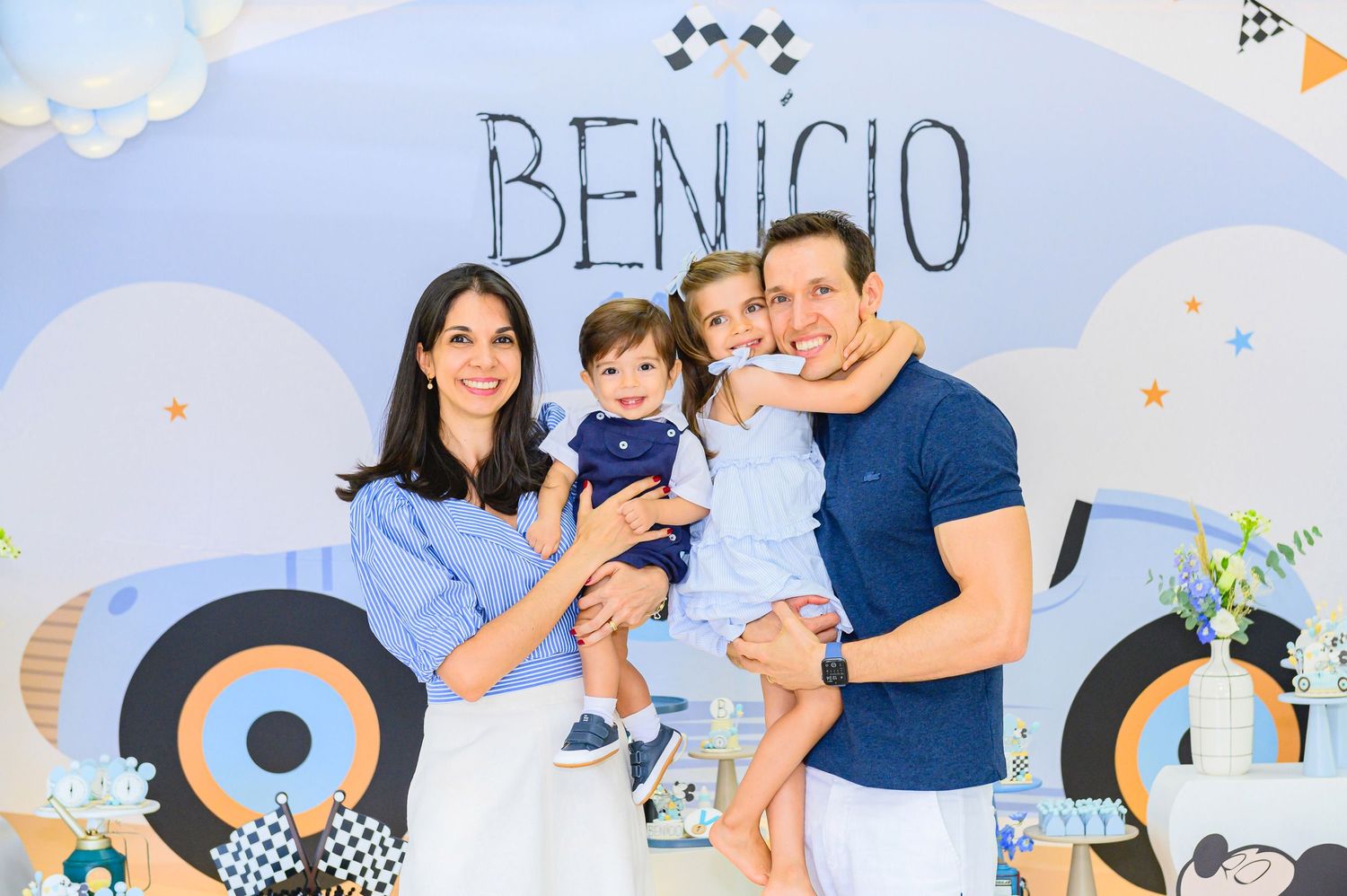 A group shares a joyful moment at a baby's 'Benicio' themed birthday party with blue and geometric wall decorations.