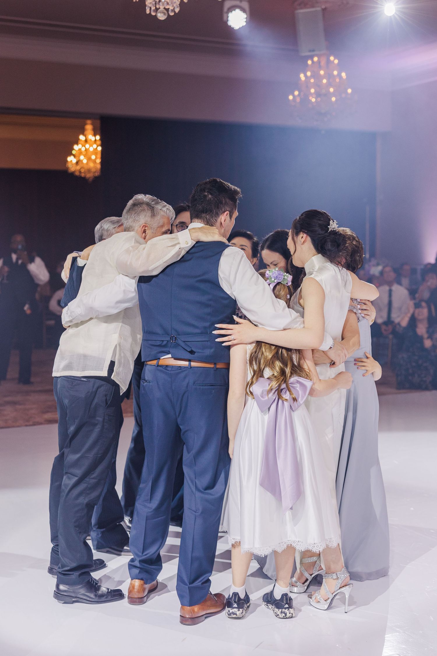 Wedding party group hug on dance floor at elegant reception venue with chandelier lighting.