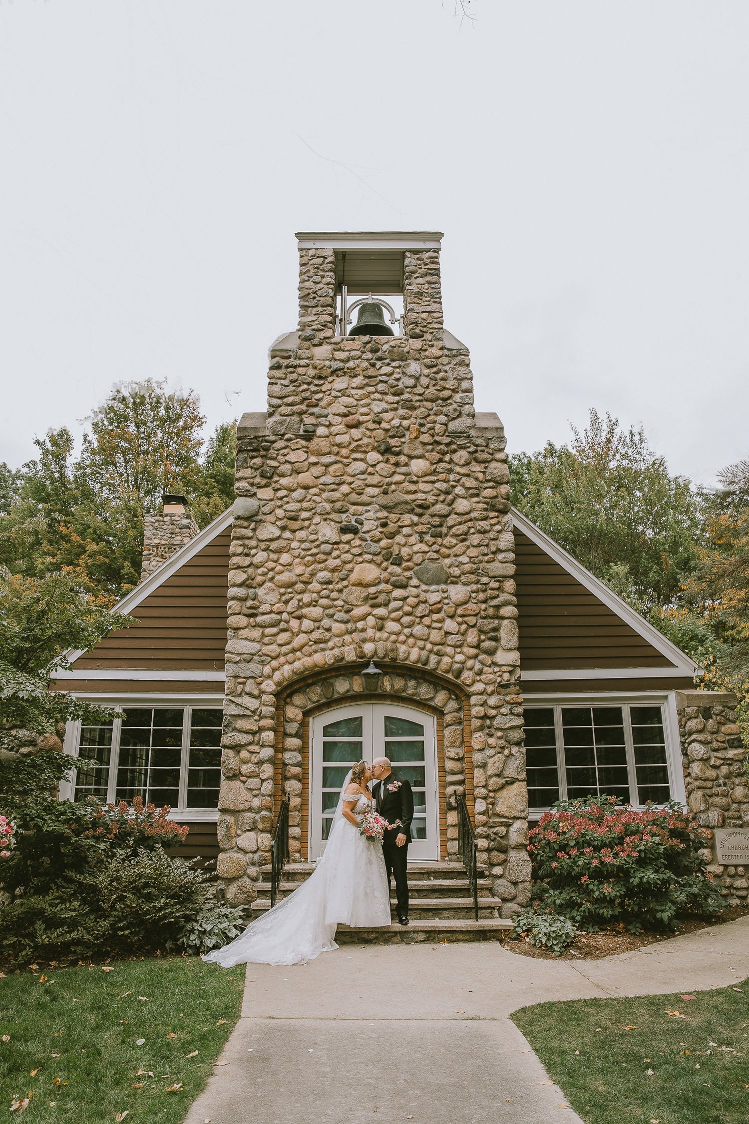 Bride and groom pose before a charming stone chapel with bell tower, surrounded by lush greenery on their wedding day.