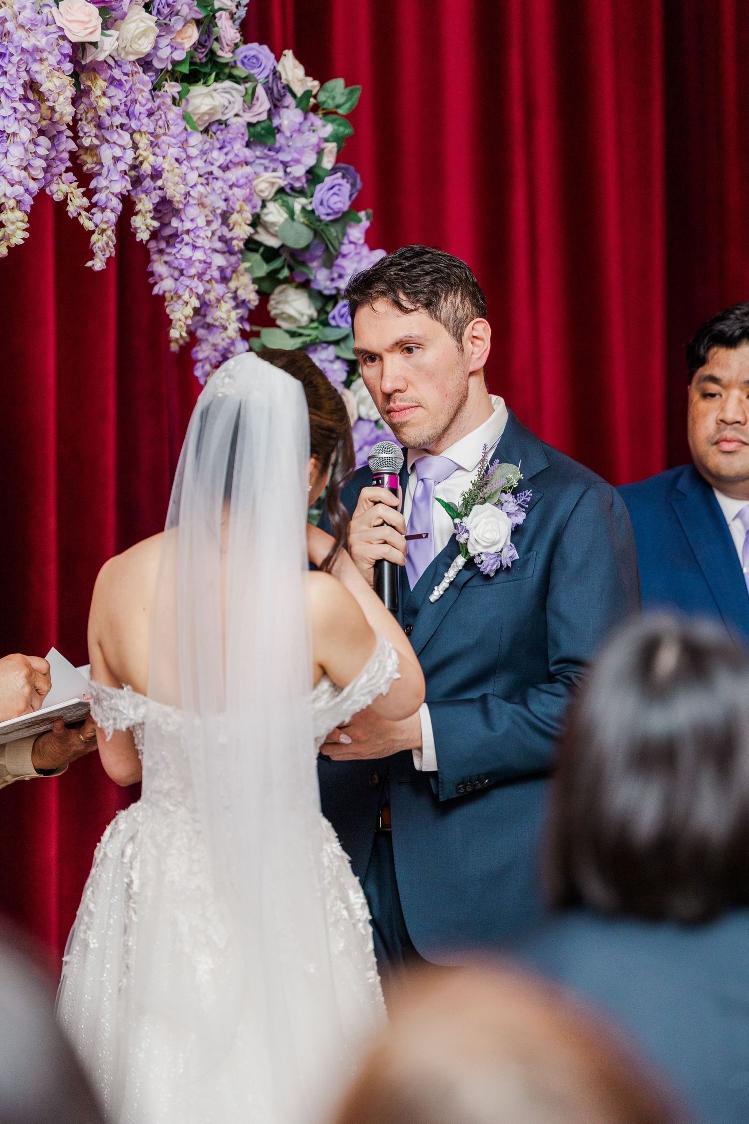 Groom in navy suit reciting wedding vows at altar adorned with purple flowers before red curtain backdrop.