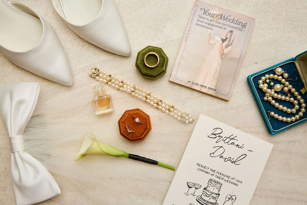 Wedding flat lay with bridal shoes, pearl jewelry, ring box, perfume, and invitation.