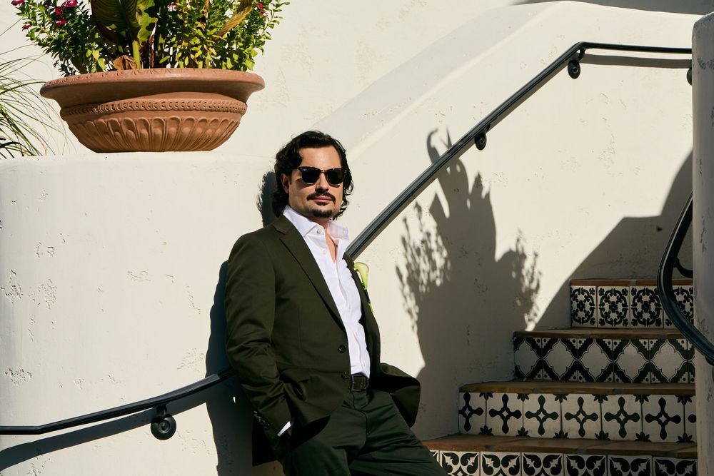 Man in dark green suit and sunglasses poses on tiled steps beside white wall with hanging terracotta planter.
