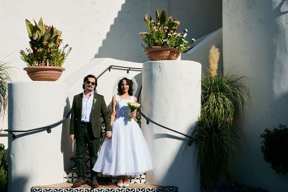 Bride and groom pose together on steps outside a white stucco building with tropical plants.
