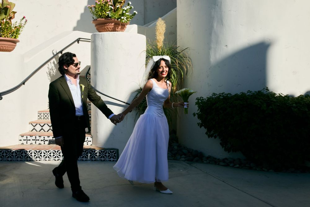 Bride in blue tulle dress and groom in dark suit hold hands outdoors at a sunny wedding venue.
