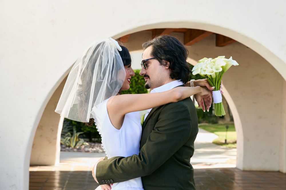 Bride and groom embrace under an arched doorway, bride holding white calla lily bouquet in outdoor wedding setting.