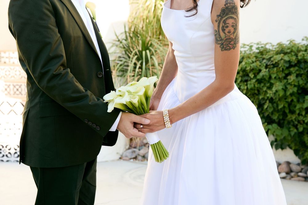 Bride and groom holding white calla lily bouquet at wedding ceremony, bride wearing white dress with tattoo.