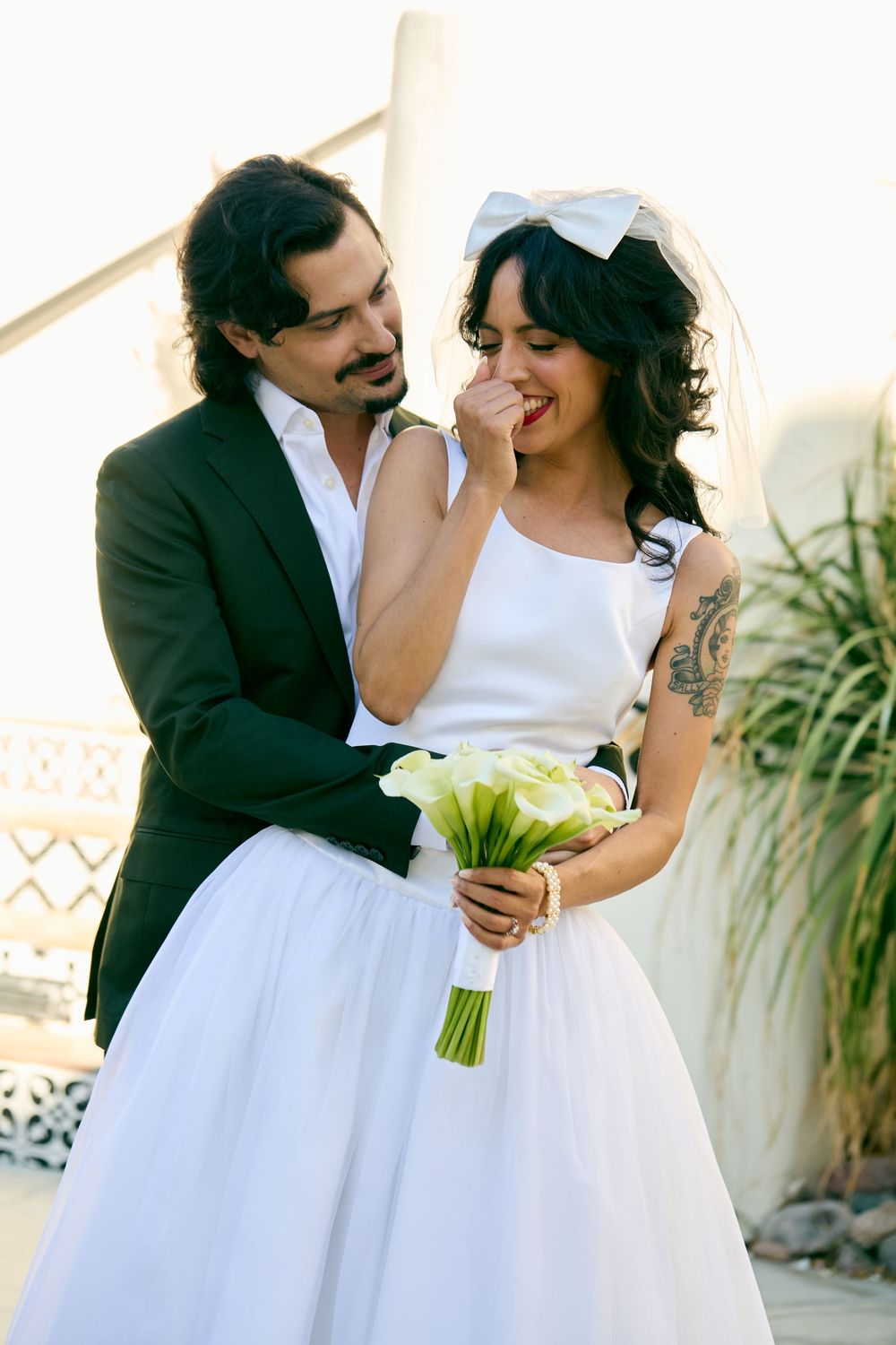 Groom in dark suit embraces tattooed bride holding white calla lily bouquet with bow veil.