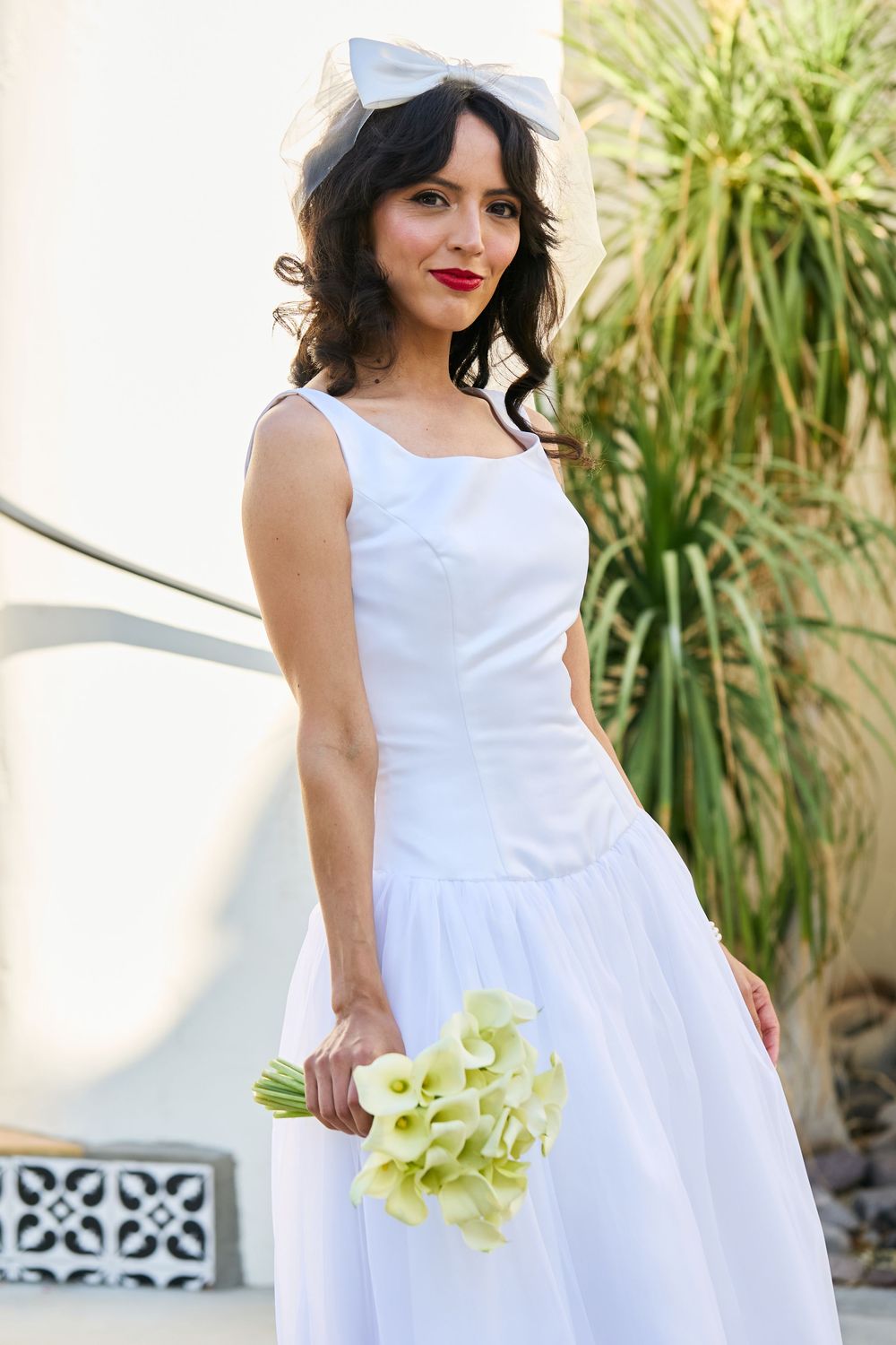 Bride in white dress holding calla lily bouquet with bow headpiece outdoors.
