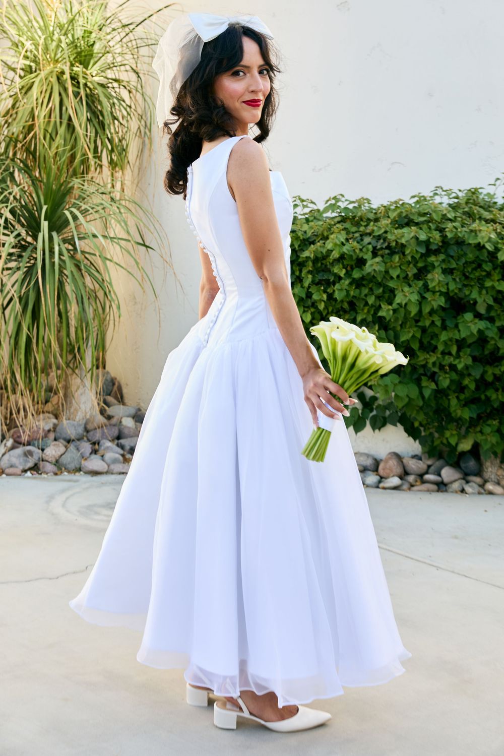 Bride in white tea-length wedding dress holding calla lily bouquet, smiling over shoulder, wearing veil and heels outdoors.