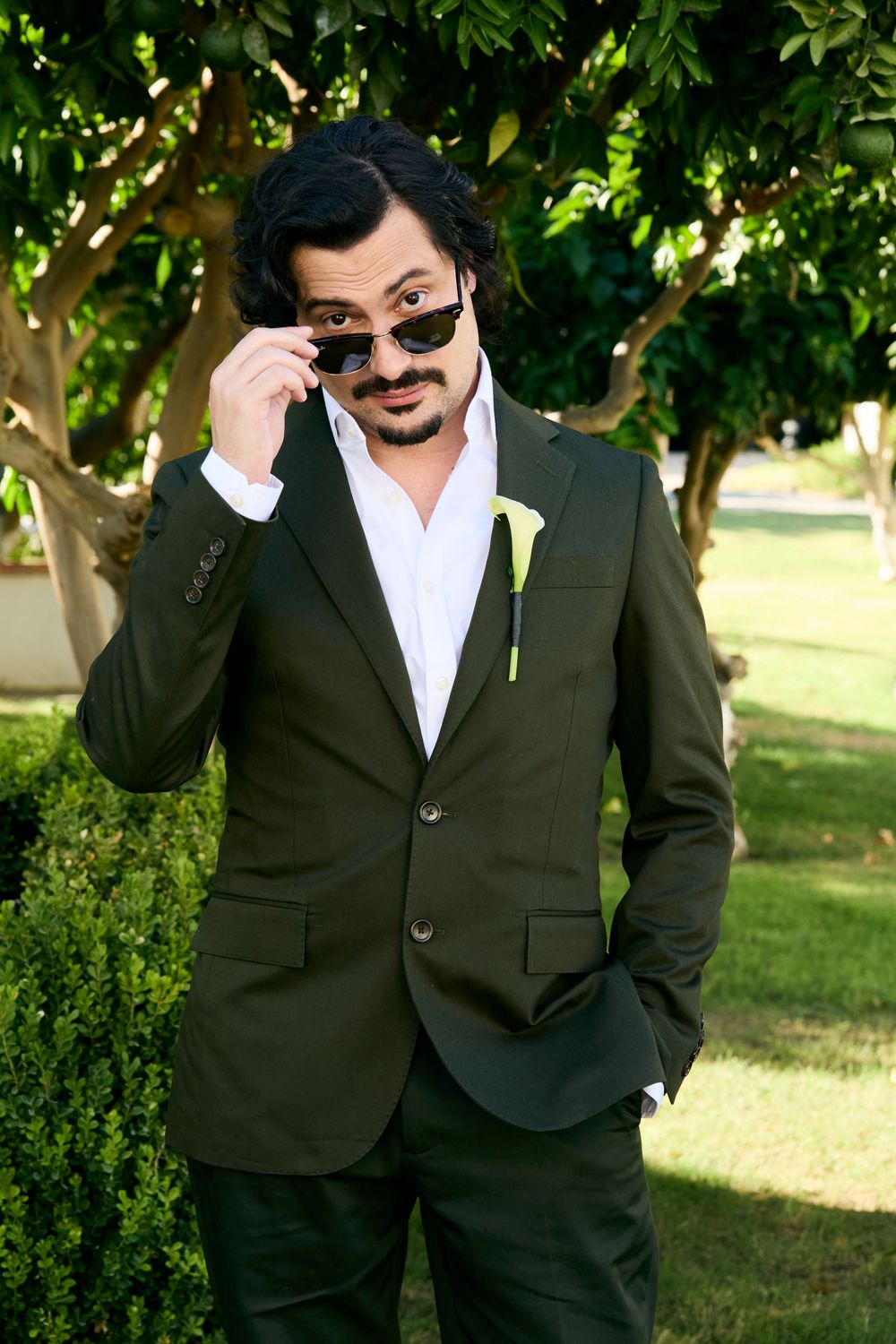 Stylish groom in dark suit with boutonniere lowering sunglasses outdoors among green trees.