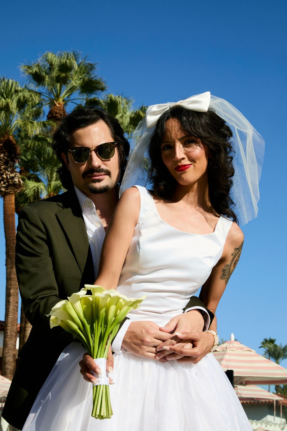 Stylish couple on wedding day, groom in dark suit with sunglasses, bride in white dress with bow veil holding calla lilies.