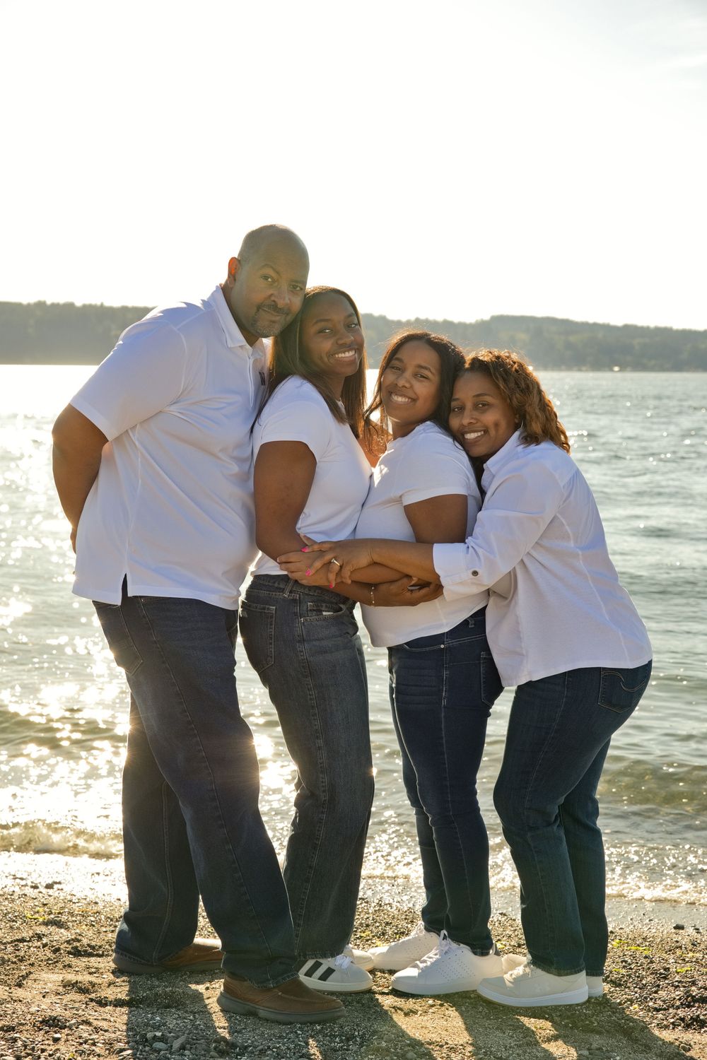 Happy family of four in white shirts and jeans posing together on a sunny beach with sparkling water in the background.