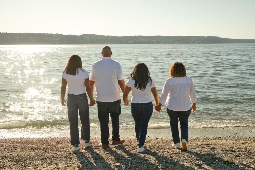 Family of four holding hands walking along a sunny beach shoreline wearing matching white shirts.
