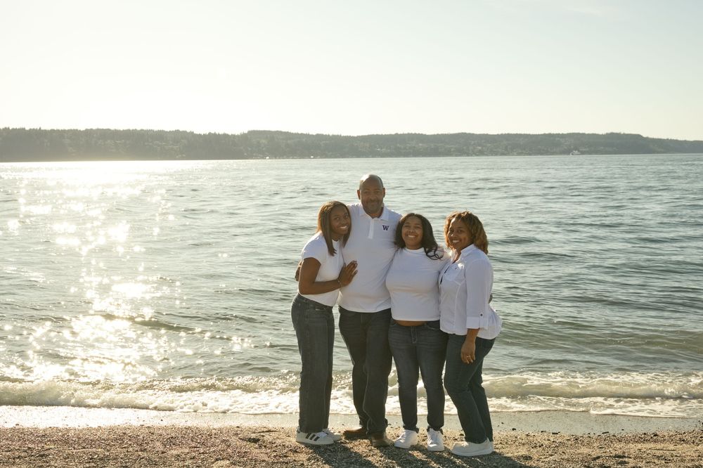 Four people posing together on a sunny beach with calm water and hills in the background.