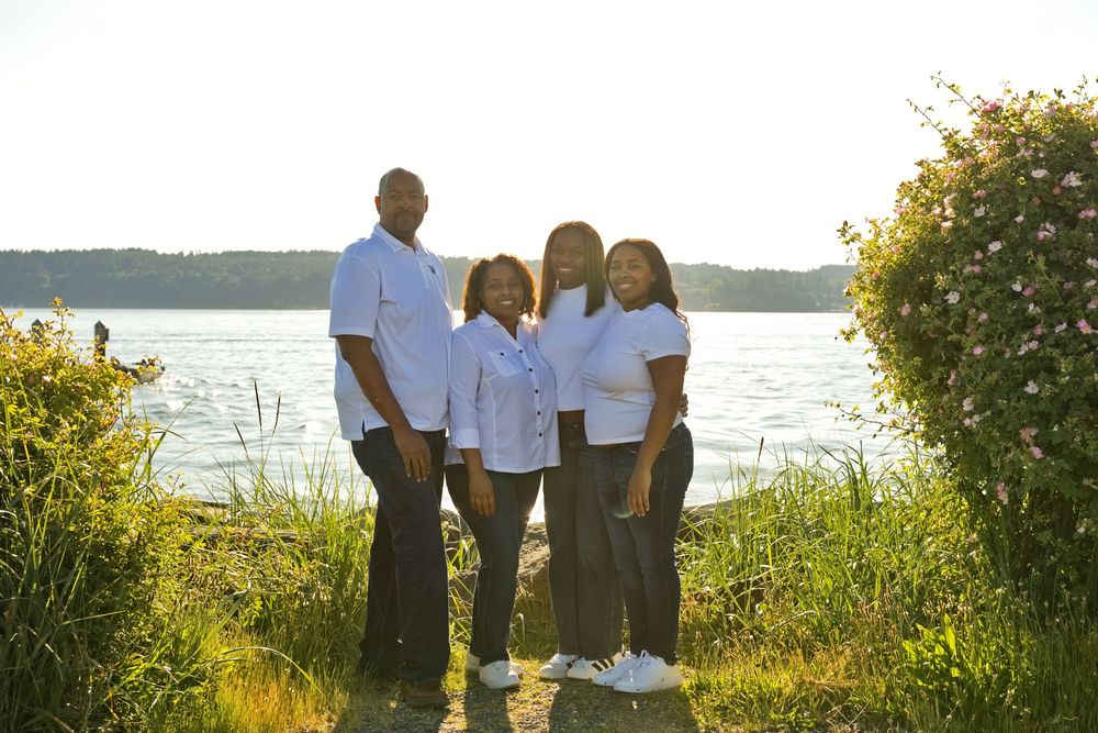 Family of four posing together near a sunlit lakeside with lush greenery and pink flowers in background.