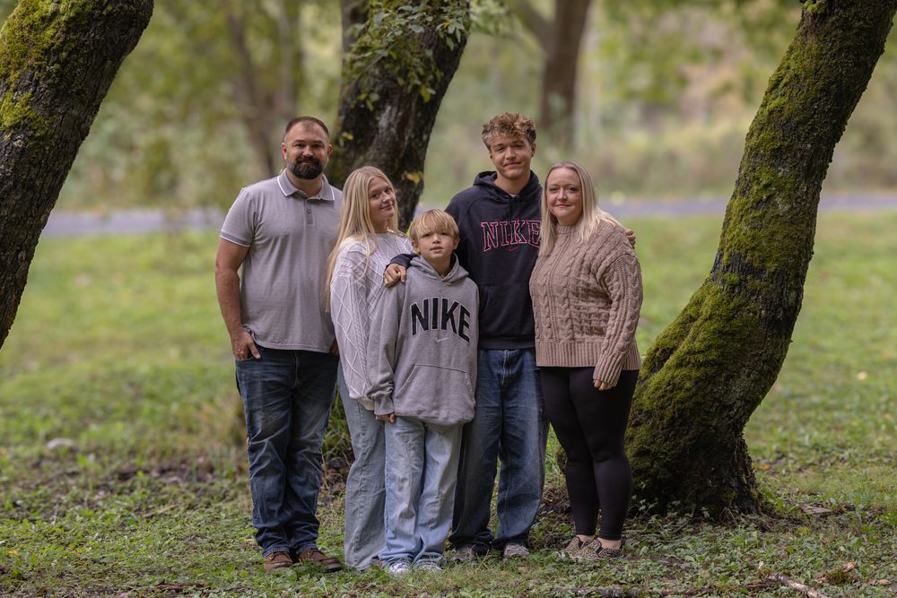 Family of five posing together outdoors among moss-covered trees in a lush green park setting.