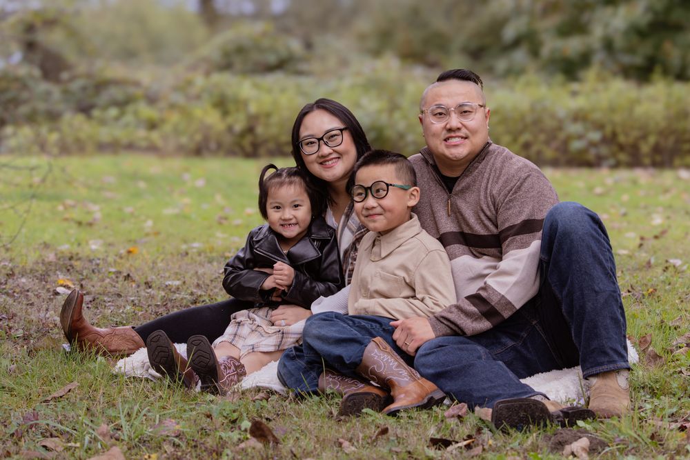 Asian family of four smiling outdoors on grass, parents with two young children wearing glasses in a park setting.