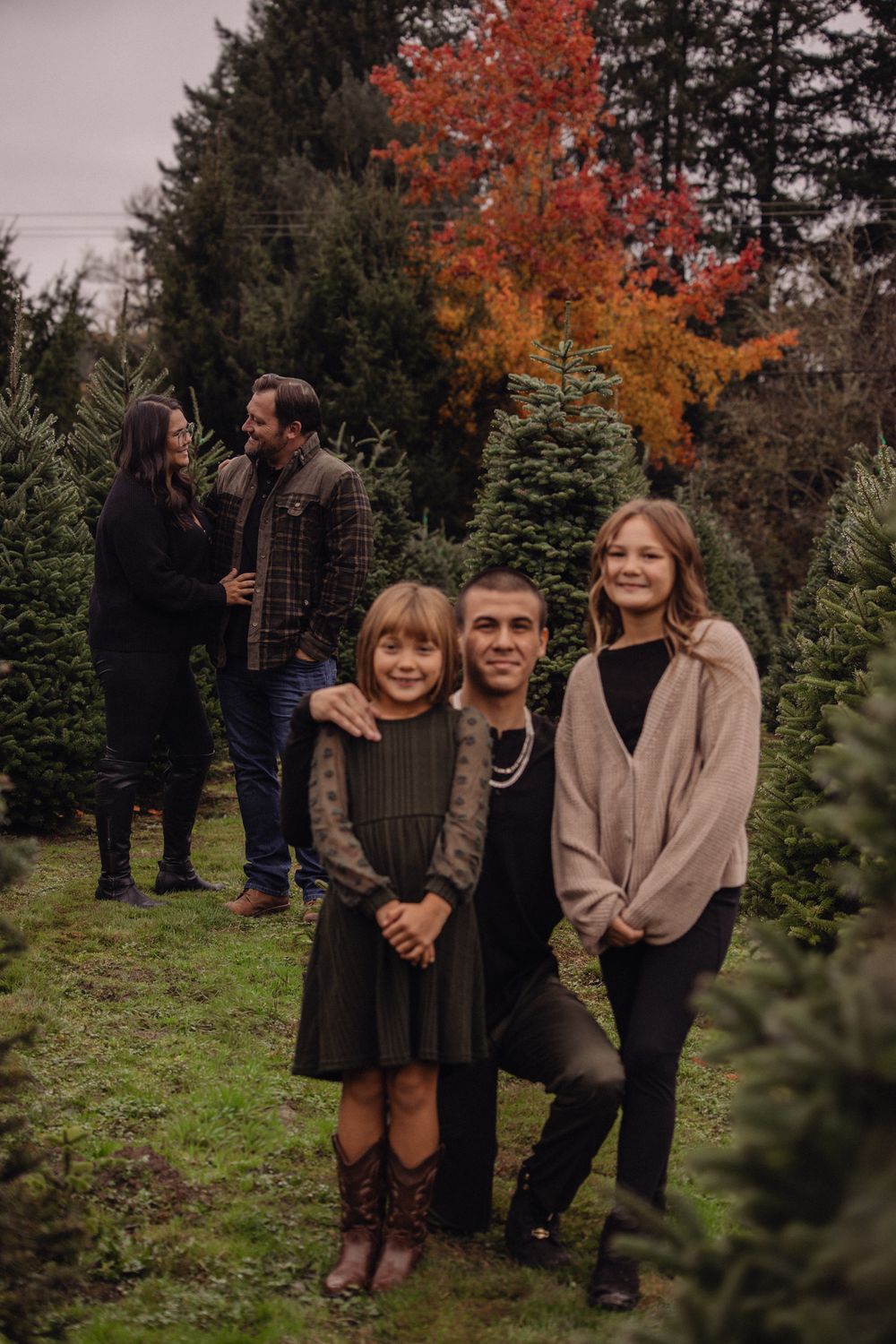 Family posing at a Christmas tree farm in autumn, with colorful foliage and evergreen trees in the background.