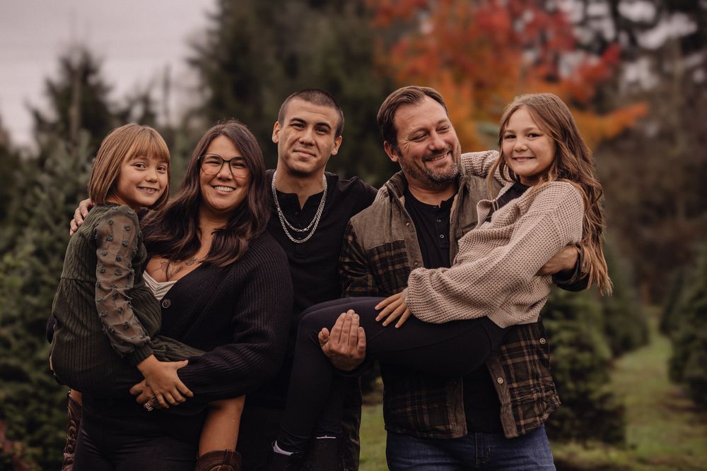 Family of five smiling outdoors at a Christmas tree farm in autumn, with colorful foliage in the background.