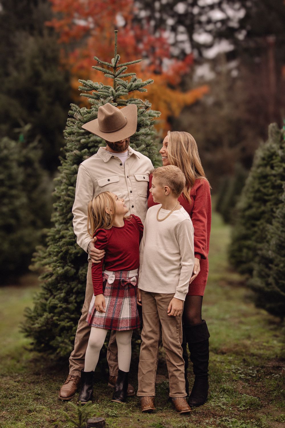 Family of four posing together at a Christmas tree farm surrounded by evergreen trees in autumn.