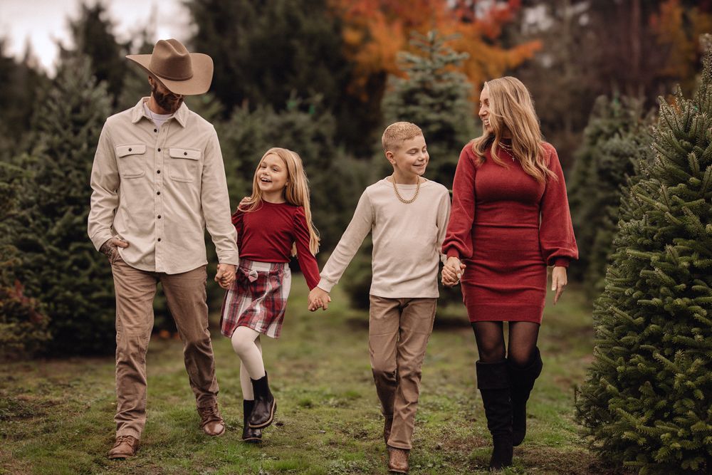 Family of four walking happily through a Christmas tree farm, dressed in festive red and tan outfits, holding hands.