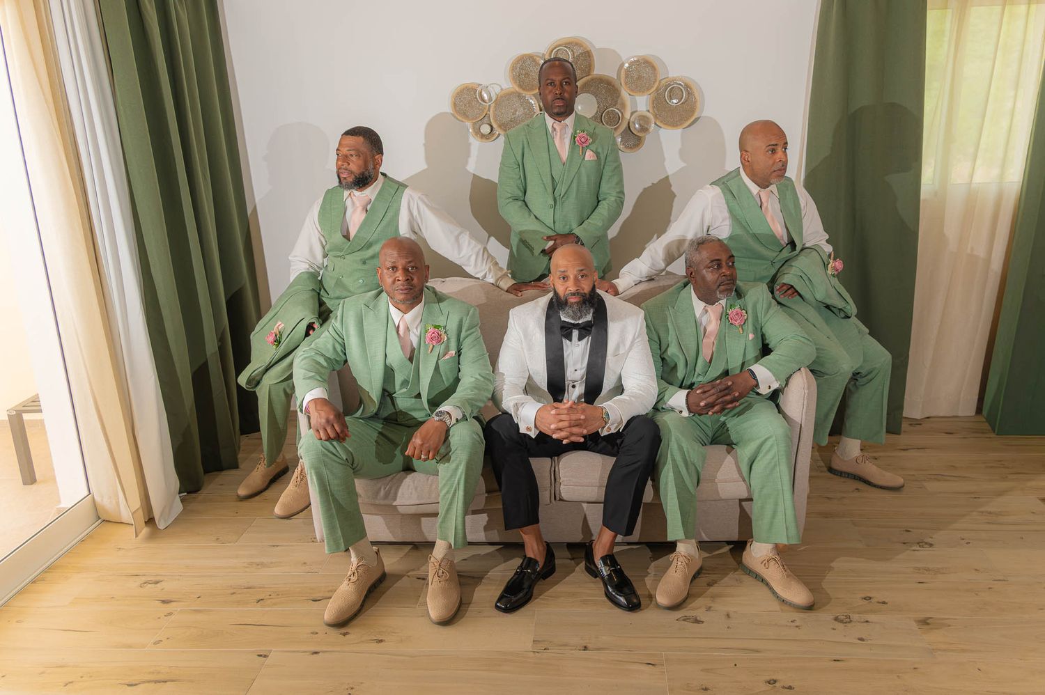 Six men in matching mint green suits pose formally, with one man in a white tuxedo seated centrally in an elegant room.