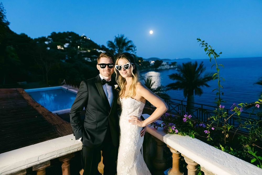 Couple in wedding attire wearing sunglasses pose on balcony overlooking ocean at night with full moon.