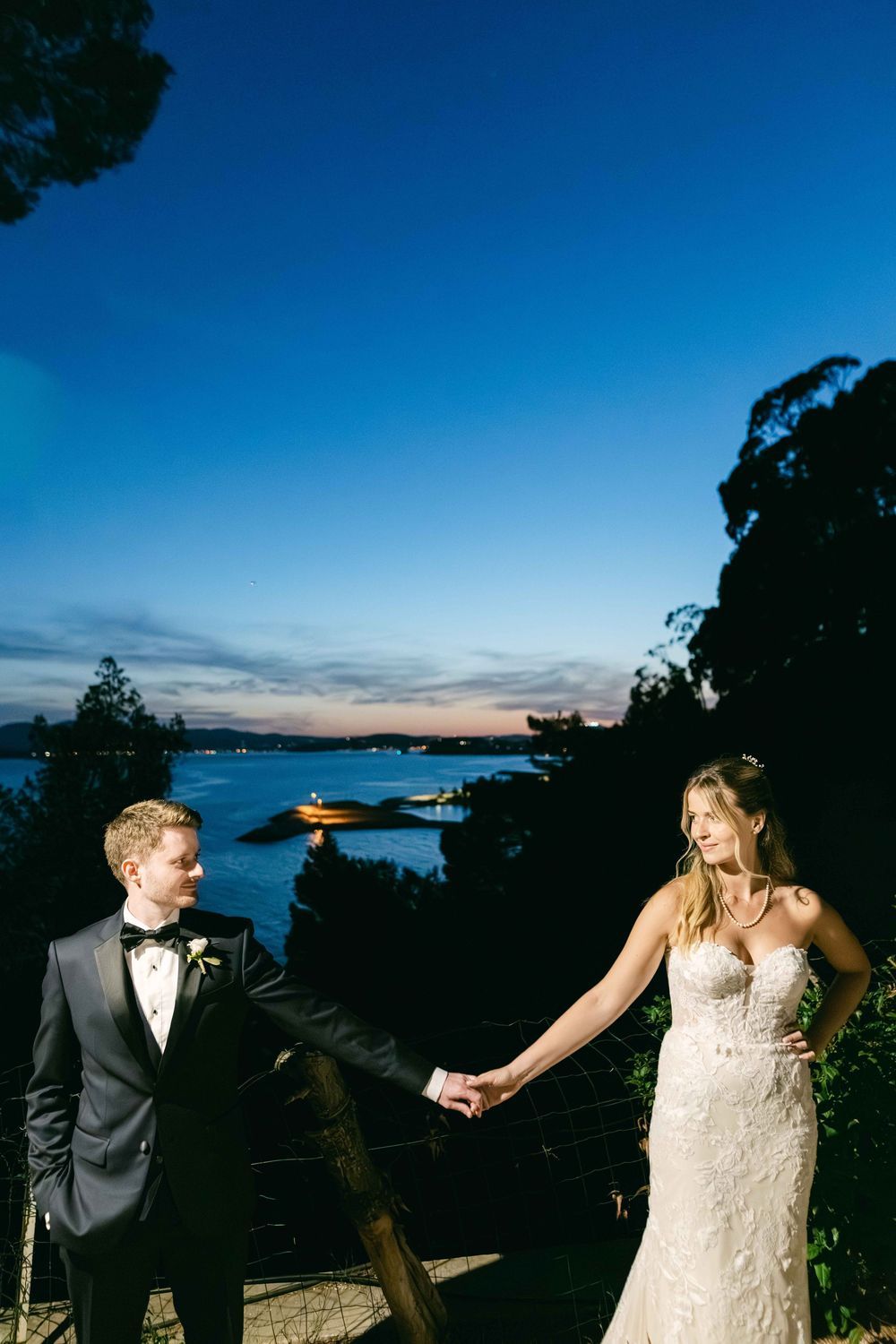 Bride and groom hold hands at twilight with waterfront views behind them, groom in tuxedo, bride in lace gown.