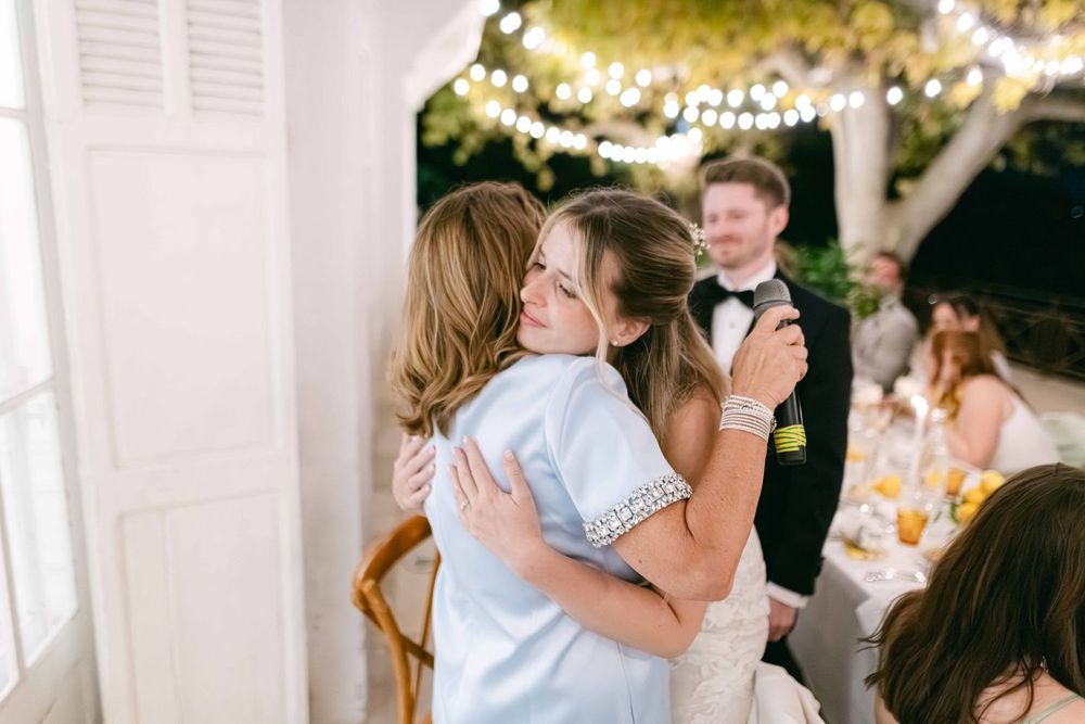 Two women share a warm hug at an elegant outdoor wedding reception with string lights overhead.