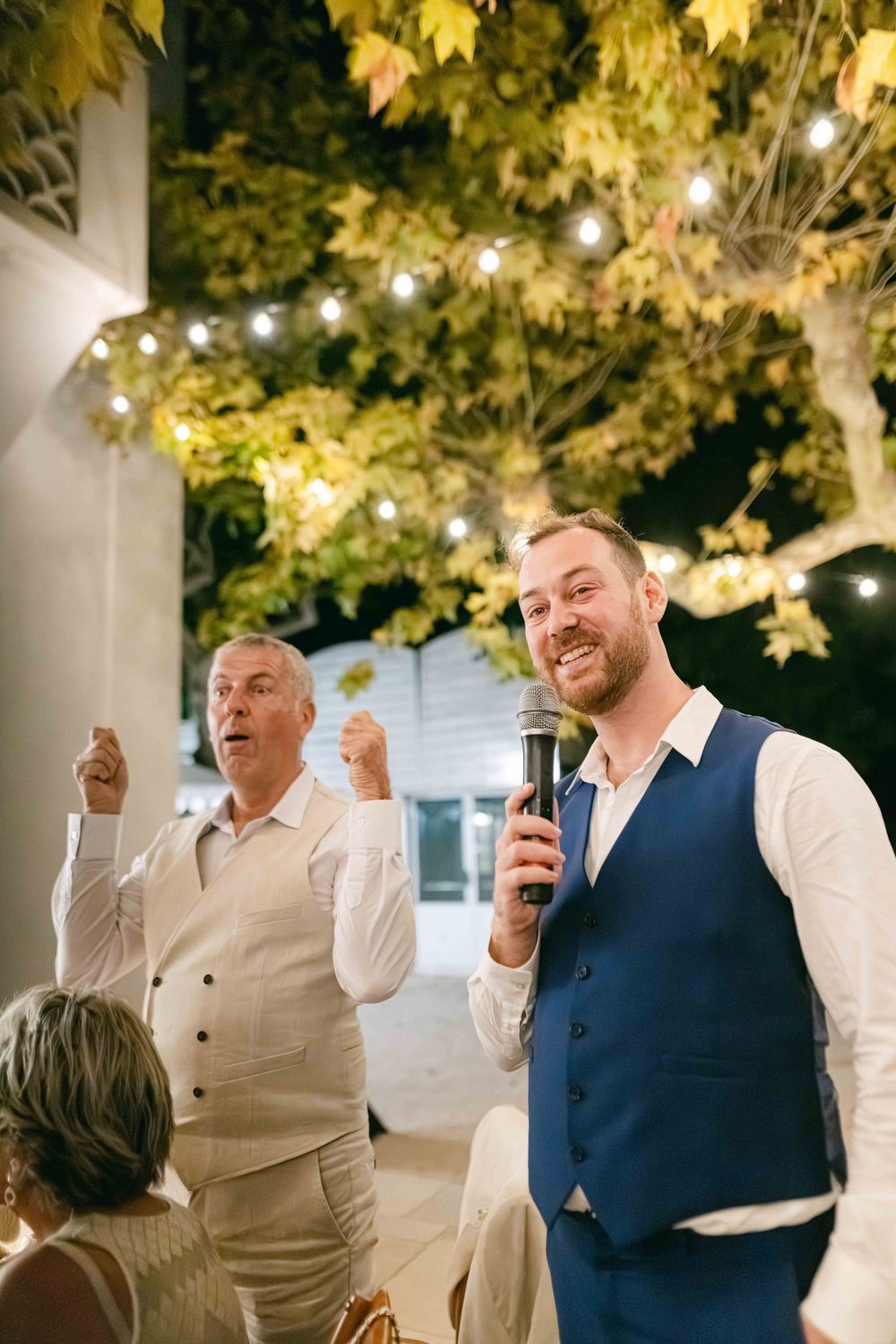 Man in blue vest gives wedding speech with microphone while older guest celebrates behind him outdoors at night.