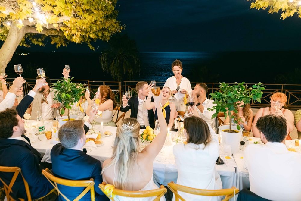 Outdoor wedding reception toast at night with guests raising glasses around elegant table with floral centerpieces.