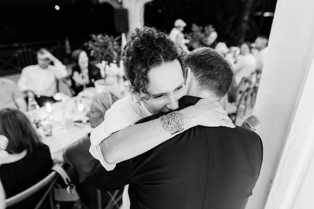 Two men share an emotional hug at a wedding reception in this black and white candid photo.