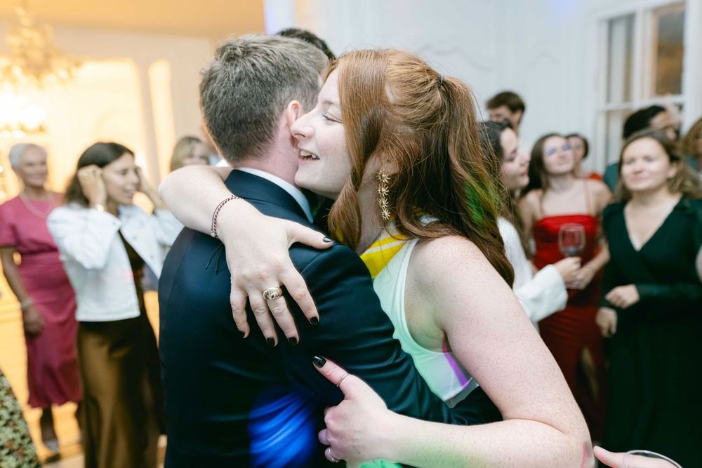 Couple embracing and smiling on a dance floor at a formal event with guests in elegant attire.